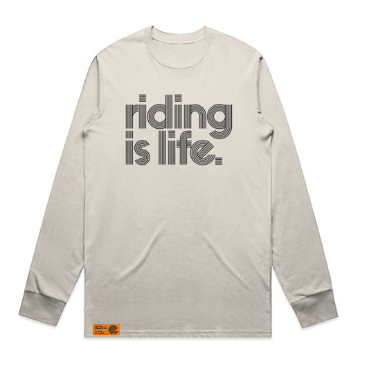 A light beige long-sleeve t-shirt with 'riding is life.' printed in a gray retro font on the chest, featuring a small orange brand label near the bottom hem.