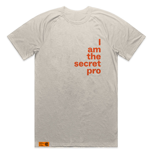 A light gray heathered short-sleeve raglan t-shirt with orange text on the chest reading 'I am the secret pro' and a small orange brand patch near the bottom left hem.