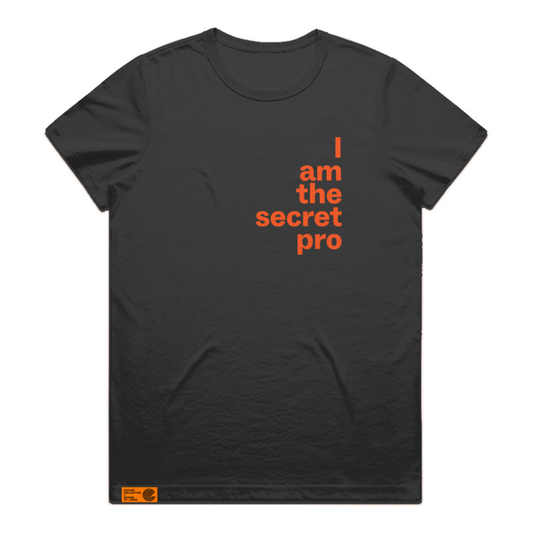 A black short-sleeve crewneck t-shirt with orange text on the right chest reading 'I am the secret pro' in a stacked, typewriter-style font. A small orange brand tag is visible near the bottom left hem.