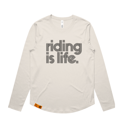A light beige long-sleeve t-shirt with 'riding is life.' printed in a bold, retro-style gray font on the chest, displayed flat against a white background.